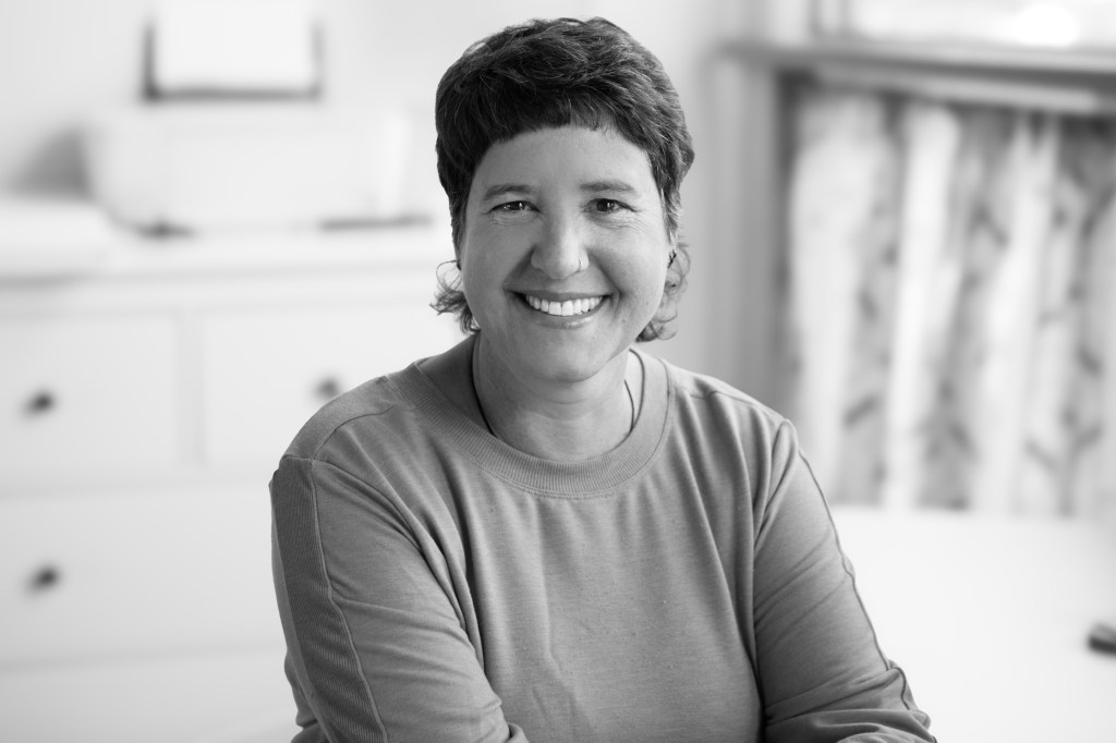 The image is Helena sitting and smiling while looking at the camera. She has short brown hair, a nose piercing and a couple of ear piercings. She is wearing a sweater. 